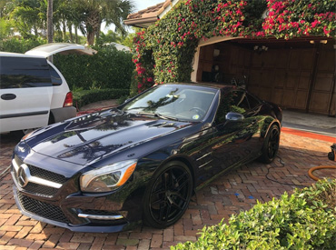 A stunning black Mercedes Benz parked in an upscale driveway surrounded by lush greenery and vibrant flowers, representing the high-quality car detailing services offered by Broward Detailers in South Florida.